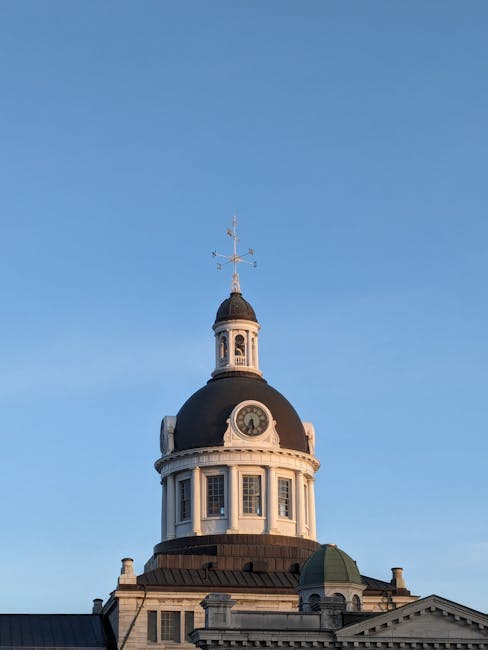 Photograph of a historic building's tower structure under a clear blue sky, featuring a black dome with a white clock face, surrounded by multiple narrow windows. The tower includes a weather vane at the top and a smaller green dome beneath the main dome. The building's stone facade shows detailed architectural features and decorative elements, indicating a classic or neoclassical style. The image captures part of the roof and upper sections of the structure, which may be part of a town hall or similar civic building in Kingston, relevant to home relocation and furniture transport services provided by Kingston Man and Van during moving and packing processes.