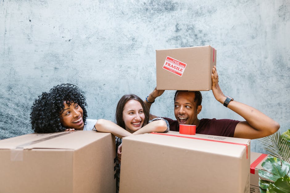 Three individuals are engaged in a home relocation process, surrounded by large cardboard moving boxes with packing tape, some labeled as fragile. The person on the left, an African American woman with curly black hair, is smiling and leaning on a box. The woman in the middle, with straight brown hair, is also smiling and resting her arms on the boxes. The person on the right, a man with short dark hair, is holding a medium-sized cardboard box with a red 'FRAGILE' sticker above his head, preparing to place it onto a stack. They are inside a room with a textured light blue wall, and natural light illuminates the scene. The environment suggests the loading and packing stage of a household move, with visible packaging materials and a focus on careful handling of fragile items, illustrating aspects of furniture transport, packing, and moving logistics handled by Kingston Man and Van.
