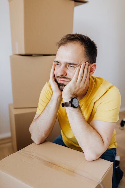 A man with short brown hair and a beard, wearing a yellow T-shirt and a black wristwatch, rests his face in his hands with a look of frustration or concern. He is seated at a light-colored wooden table in a room with plain beige and white walls. In front of him, there is a cardboard box, and behind him, stacked cardboard moving boxes are visible. The setting appears to be part of a home relocation or packing process, with the man possibly experiencing stress related to house removals or moving logistics. The lighting is natural, highlighting the emotional weight of the moment, and the scene is representative of the challenges faced during home packing or the waiting periods involved in house removals, as addressed by Kingston Man and Van's removal services.