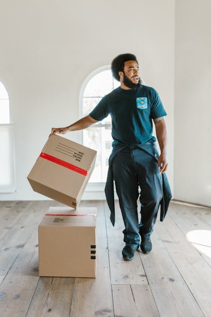 A man with dark curly hair and a beard, dressed in a navy blue t-shirt with a blue logo on the chest and navy trousers, is inside a bright, unfurnished room with natural daylight coming through a larg