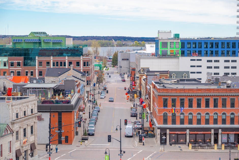 An aerial view of a town centre street during daylight, with a variety of brick and modern glass-fronted buildings lining both sides. Several parked cars are visible along the pavement and in the on-street parking bays. The street features a loading area where a large, open-top moving van is parked, with packed cardboard boxes, wrapped furniture, and plastic containers being loaded or unloaded by movers using trolleys and straps. The furniture includes large wardrobes and medium-sized boxes, with some wrapped in protective blankets. A few movers can be seen handling items, coordinating the loading process inside and outside the van. The scene captures the logistics of home relocation, with a focus on furniture transport, packing, and loading procedures, while also referencing Kingston council rules for parking suspensions during removals, as indicated by the surroundings and signage visible on the street.