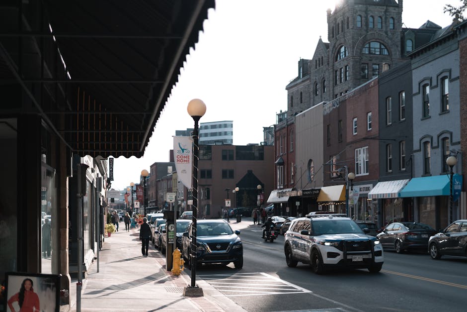 A street scene in Kingston showing a mix of parked and moving vehicles including a police car and a black SUV, with shops and buildings lining the road. The pavement features pedestrians walking and some carrying items, while parked cars are positioned along the curbside. Streetlights and signage are visible, and the overall environment suggests a busy urban area, consistent with a home relocation or furniture transport process. Kingston Man and Van may be involved in the scene as part of their removals service, supporting logistics for local house moves. The lighting indicates daytime, with a clear sky overhead, and the street appears to be within a commercial district with various small businesses and storefronts.