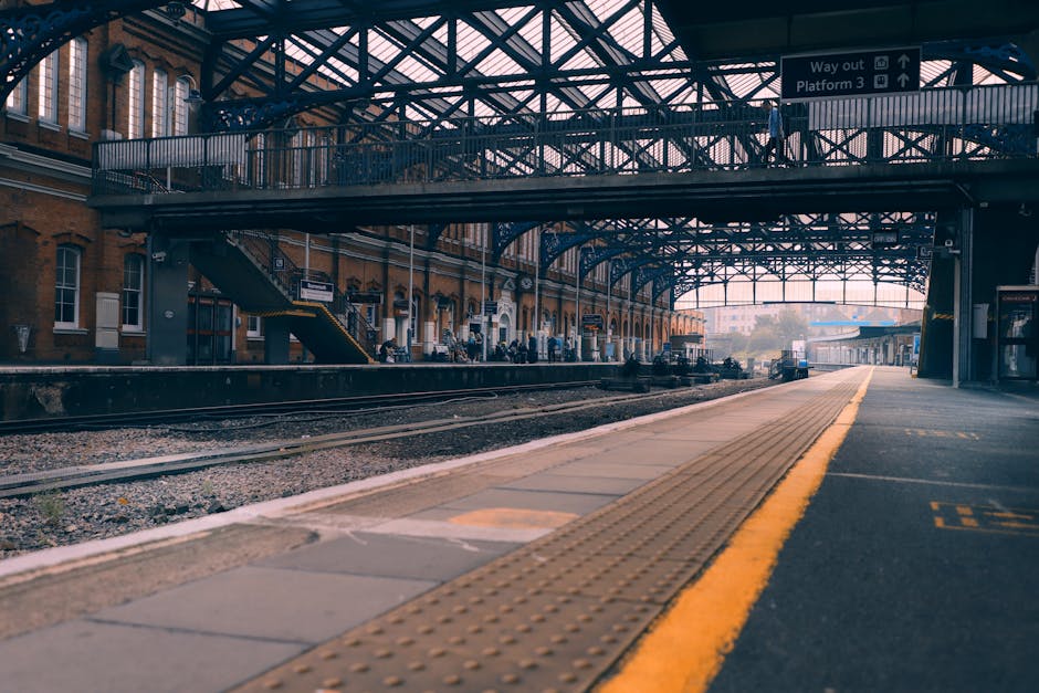 A wide-angle view of Kingston Station platform showing a partial covered structure with a decorative iron framework and glass panels overhead. The platform surface is paved with concrete and features tactile paving strips along the edge for accessibility. In the midground, several cardboard-packed boxes and small furniture pieces are being loaded onto a blue moving van parked adjacent to the platform. The van’s rear doors are open, and moving straps and blankets are visible, indicating a home relocation process. Nearby, a trolley and other packing materials support the furniture transport transition. The station’s historic brick building, with multiple windows and a staircase, is visible along the platform, with a few commuters waiting or walking. Overhead, signage points toward exits, platforms, and routes, supporting efficient logistics for a move. The environment is illuminated by natural daylight, emphasizing the busy, organized loading process commonly associated with professional house removals and moving services like Kingston Man and Van.