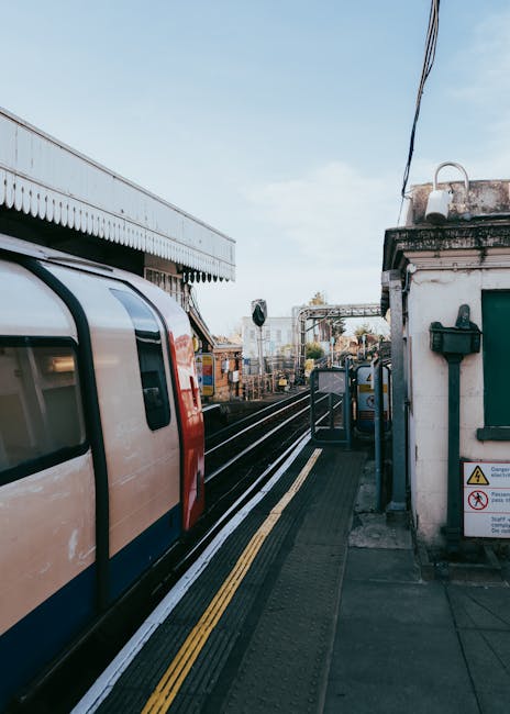 The image shows a view of Kingston Station platform during daytime, with a train stationary on the tracks next to the platform edge. The train has a modern design with a cream and red exterior, and visible windows along its side. The platform has a textured, slip-resistant surface with yellow tactile paving strips near the edge for safety. On the right side, there is a small white building with a green window and a black mailbox attached to the wall, along with some signage indicating safety instructions. Overhead power lines and cables run above the platform, with a clear blue sky in the background, suggesting good weather. The station appears to be quiet with no passengers visible in the scene. This setting illustrates transportation infrastructure relevant for home relocation or furniture transport, as part of the moving process managed by Kingston Man and Van, specializing in removals and moving services in the Kingston area.