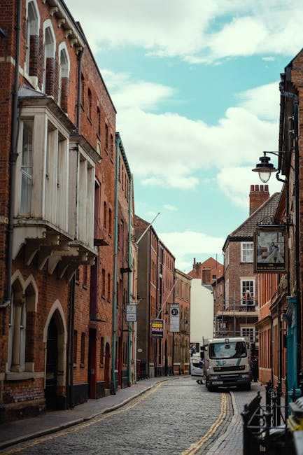 A narrow urban street scene during daytime shows brick residential and commercial buildings on both sides, with some featuring traditional architectural details such as bay windows and decorative brickwork. The street is paved with cobblestones and has a double yellow line along the curb. A white van from Kingston Man and Van is parked near the middle of the street, with its rear doors open, revealing a loading area where furniture and boxes are visible being carefully loaded or unloaded. Nearby, a hand truck or trolley is positioned next to the van, ready for use. The open doors of the building on the left reveal a glimpse of packed furniture and boxes inside, indicating a home relocation or moving process. Street lamps and signage are visible mounted on the buildings, and the sky above is partly cloudy with blue patches. The scene captures the typical environment of a challenging narrow street move, with careful furniture transport and packing preparations, reflecting Kingston Man and Van's moving services for tight urban streets.