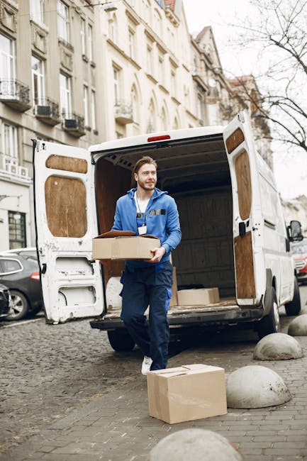 A male mover from Kingston Man and Van, dressed in a blue hoodie and work trousers, carrying a cardboard box during a home relocation process on a cobblestone street in Kingston. The van's rear doors are open, revealing additional boxes inside, some wrapped in plastic or paper. Outside the vehicle, a large cardboard box and another smaller package rest on the pavement near a pair of rounded concrete bollards. Residential buildings with cream-colored facades and traditional architecture line the background, while parked cars are visible along the street. The scene is set during daytime under overcast skies, illustrating the loading process involved in furniture transport and packing for a house move, emphasizing careful handling and organized logistics typical of professional removals by Kingston Man and Van.
