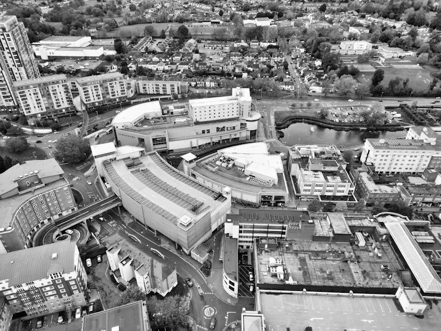An aerial black-and-white photograph showing a cityscape with a large modern building complex in the foreground, part of a home relocation or furniture transport process. Inside the complex, several cardboard boxes and plastic-wrapped furniture are visible, indicating packing and moving activities. To the left, a van belonging to Kingston Man and Van is parked on the street, with a person lifting and carrying a box towards the building's entrance. The background features residential houses, green parks, and a small pond, all situated within an urban environment. The scene captures the logistical detail of loading items for house removals, with equipment such as trolleys or straps possibly seen supporting the moving process on the pavement. The overall setting highlights professional packing and furniture transport operations as part of an organized move in Kingston near the Bentall Centre, illustrating aspects of logistics, loading, and transport related to house removals services.