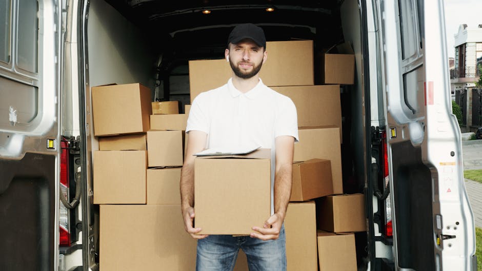 A man wearing a white polo shirt and glasses is seen loading or unloading cardboard boxes from the back of a white moving van parked on a suburban street during sunset. The van's open rear door reveal