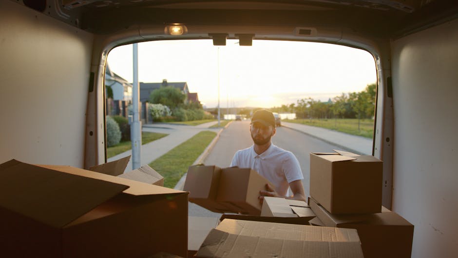 A man wearing a white polo shirt and sunglasses is standing outside a white van with an open rear door, engaged in a home relocation task during sunset. The van's interior is visible, filled with cardboard boxes of various sizes, some sealed with packing tape and others open, revealing packing materials such as bubble wrap and paper. The man is in the process of loading or unloading boxes, with several placed on the ground nearby. The scene is set on a suburban street with a sidewalk, a lamppost, and residential houses surrounded by greenery in the background. The lighting suggests late afternoon or early evening, with warm sunlight casting a glow over the scene. This image reflects professional furniture transport, packing, and loading processes associated with same-day man and van Kingston move services offered by Kingston Man and Van.