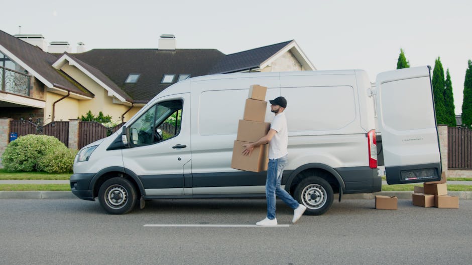 A man wearing a white t-shirt, jeans, and a black cap is loading three large cardboard boxes into the side of a white panel van parked on a residential street. The van, operated by Kingston Man and Van, is glossy and modern, with its sliding door open revealing a spacious cargo area suited for home relocation or furniture transport. The man is holding one of the boxes with both hands, positioning it on top of previously loaded boxes positioned vertically against the van's side. Additional boxes rest on the pavement beside the open sliding door, ready for the loading process. The street features neatly maintained lawns, shrubbery, and modern detached houses with pitched roofs, dormer windows, and a wooden fence separating yards. The scene is set during the daytime with natural light illuminating the surroundings, exemplifying efficient packing and loading during an urgent same-day move, with the focus on professional removals and transport logistics.